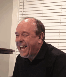 a bald man in a black shirt is smiling in front of a window with blinds