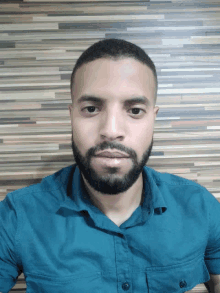 a man with a beard wearing a blue shirt is standing in front of a wooden wall