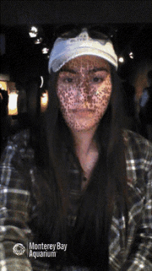 a woman with spots on her face is wearing a plaid shirt and a white hat from the monterey bay aquarium