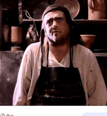 a man wearing an apron and a hat is standing in front of a shelf with pots and pans .