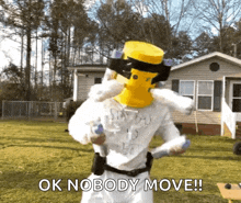 a man with a yellow hat on his head is standing in front of a house holding a gun .