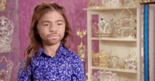 a young boy with long hair and a beard is standing in front of a shelf filled with crowns and tiaras .