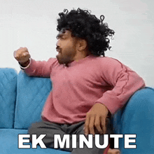 a man wearing a wig is sitting on a blue couch with the words " ek minute " written on the bottom