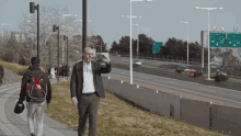 a man in a suit walks down a sidewalk near a highway with a green sign that says ' avenida '