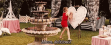 a woman in a red dress is holding a large heart in front of a fountain and saying mmmm delicious .