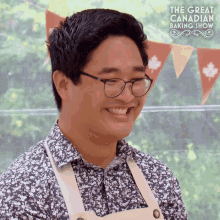 a man wearing glasses and an apron is smiling in front of a banner that says the great canadian baking show