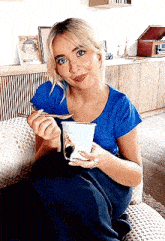 a woman in a blue shirt is sitting on a couch eating a bowl of food with a spoon .