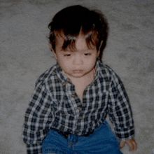 a young boy wearing a plaid shirt and jeans sits on a carpet