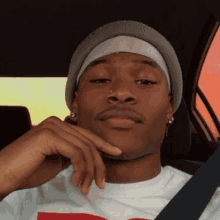 a young man wearing a beanie and a headband is sitting in a car with his hand on his chin .