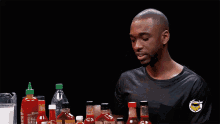 a man wearing a black shirt with a shark on it stands in front of a row of hot sauce bottles