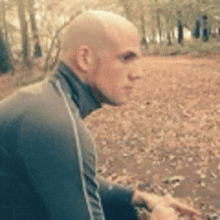 a bald man in a black jacket is standing in the woods with leaves on the ground .