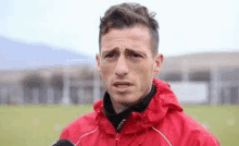 a man in a red jacket is standing on a soccer field with a microphone .