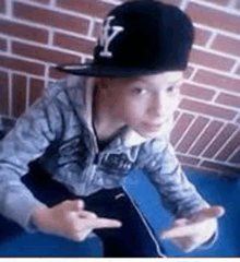 a young boy wearing a hat and a hoodie is sitting in front of a brick wall and giving the middle finger .