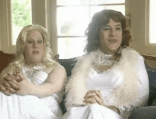 two women are sitting on a couch and one is wearing a white fur coat