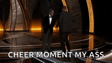 a man in a tuxedo is standing on a stage with the words cheer moment my ass behind him .