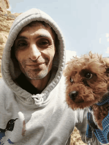 a man wearing a hooded sweatshirt with a bird on it holds a small brown dog