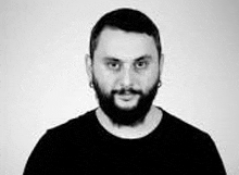 a man with a beard and earrings is wearing a black shirt and looking at the camera in a black and white photo .