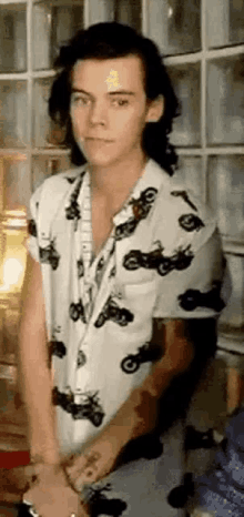 harry styles is wearing a white shirt with motorcycles on it and is standing in front of a glass wall .