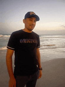 a man wearing a black shirt that says original stands on a beach