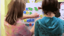 two girls are playing with magnetic letters on a white board and one of them is wearing a blue hoodie