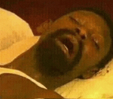 a man with a beard is laying in a bed with his mouth open .