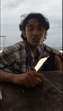 a man wearing a plaid shirt sits at a table on the beach