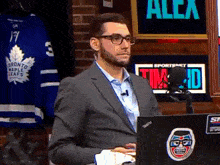 a man in a suit sitting in front of a laptop with a toronto maple leafs jersey in the background