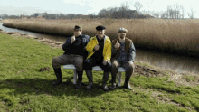 three men are sitting on a bench in a grassy field and one of them is wearing a yellow jacket