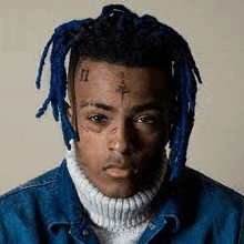 a man with blue dreadlocks and a tattoo on his face is wearing a turtleneck sweater and a denim jacket .