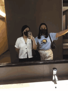 two women wearing face masks are taking a selfie in a bathroom mirror