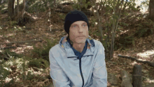 a man wearing a black beanie and a white jacket is sitting in the woods