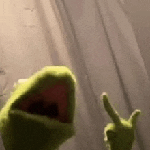 a close up of a kermit the frog puppet giving a thumbs up in front of a curtain .