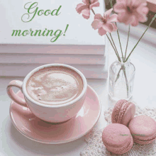 a cup of coffee sits on a saucer next to pink macaroons and a vase of pink flowers