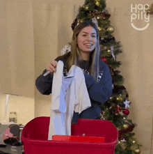 a woman is holding a white shirt in front of a christmas tree and the word happy pill is on the wall behind her