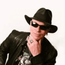 a man wearing a hat , sunglasses , and a leather jacket is holding his hand to his chin .