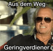 a man wearing sunglasses and a black shirt with the words `` aus dem weg geringverdiener '' written on it .