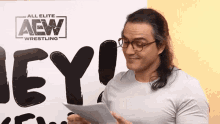 a man wearing glasses holds a piece of paper in front of a sign that says aew wrestling