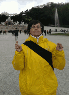 a woman wearing a yellow jacket and glasses stands in front of a fountain