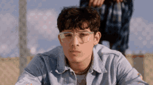 a young man wearing glasses and a denim jacket is looking at the camera