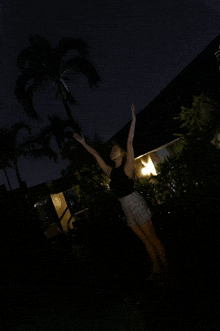 a woman in a black tank top and plaid shorts stands in the dark with her arms outstretched