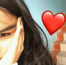 a close up of a woman covering her face with her hand and a red heart emoji behind her .