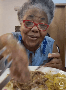 a woman with glasses is eating a plate of food with a likes icon on the bottom right