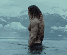 a man without a shirt is standing in the water with mountains in the background .