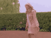 a woman in a pink suit and hat is standing in front of a bush