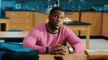 a man in a pink sweater is sitting at a table with his hands folded .