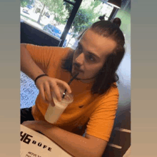 a young man with long hair is sitting at a table drinking a milkshake with a straw .