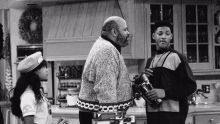 a black and white photo of three people in a kitchen one of whom is wearing a beret with the word army on it