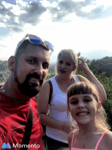 a man in a red shirt is taking a selfie with a woman and a girl with the words momento on the bottom right