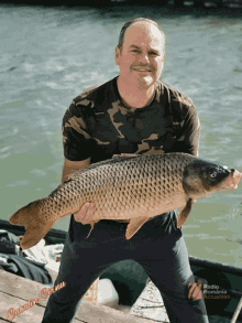 a man in a camo shirt holds a large carp in front of a sign that says radio romania actualitati