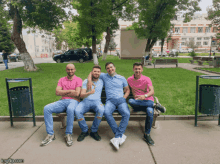 four men are sitting on a park bench with their arms crossed and a green trash can in the foreground